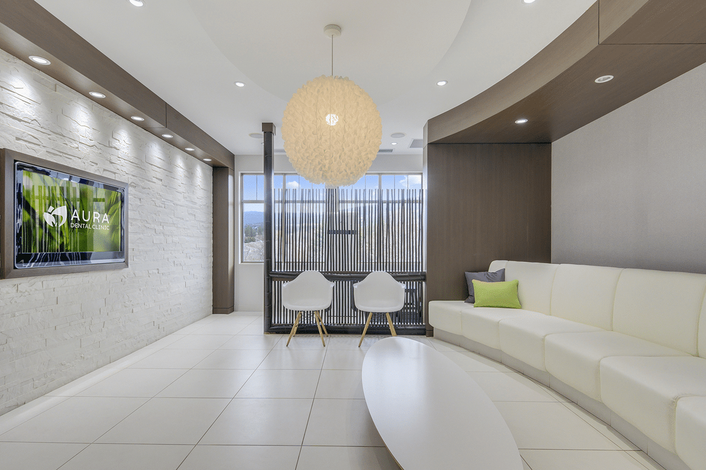 Modern dental clinic waiting room with a white couch, two chairs, a round light fixture, and a TV displaying the clinic's logo.