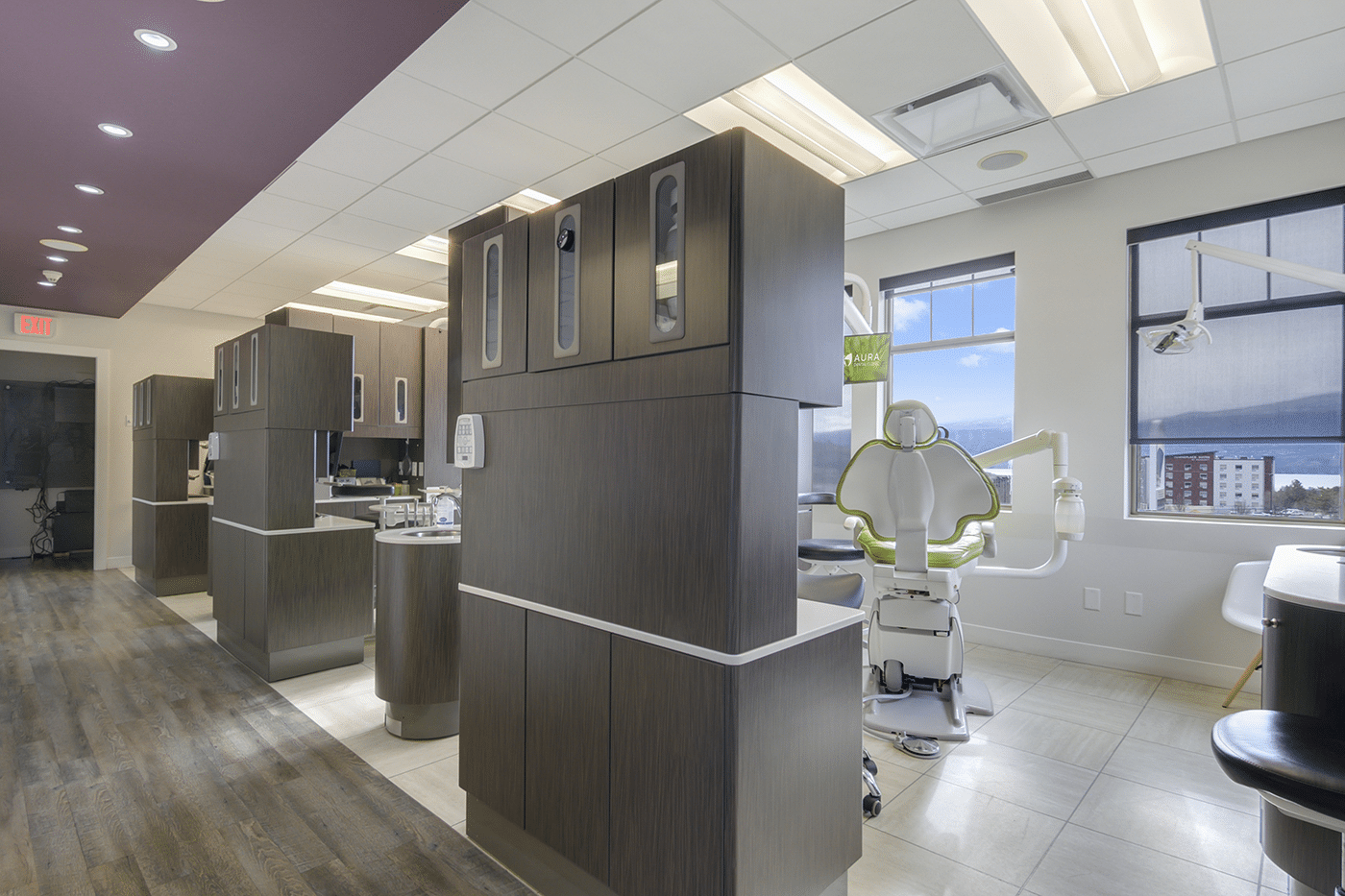 A modern dental clinic interior featuring treatment stations with storage cabinets, a dental chair, and large windows.