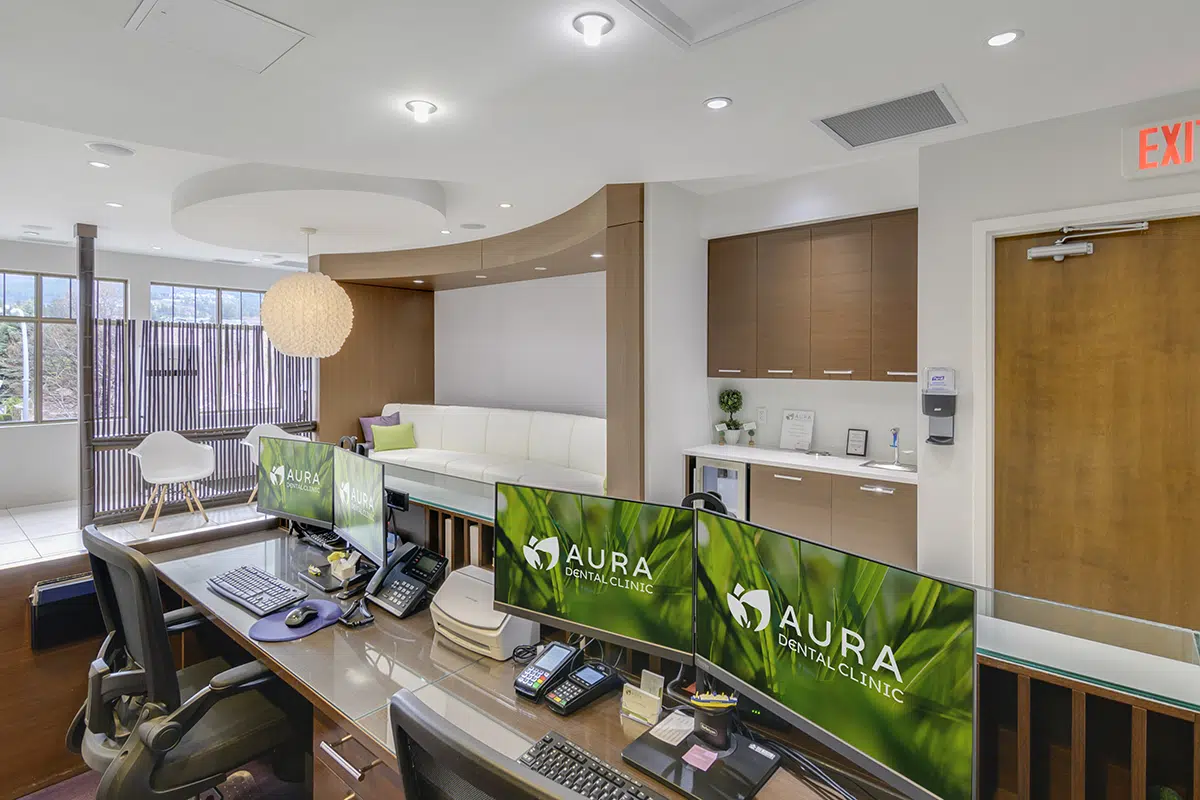 A modern dental clinic reception area featuring two computer monitors, phones, and a welcoming seating area with a couch and a decorative lamp.