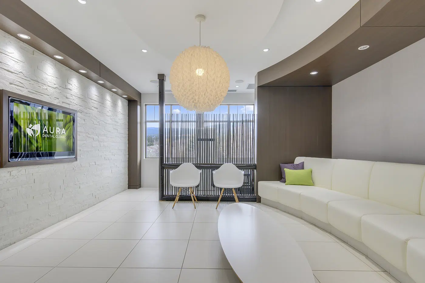 A modern dental clinic waiting area featuring a white curved sofa, two white chairs, a round light fixture, a wall-mounted TV