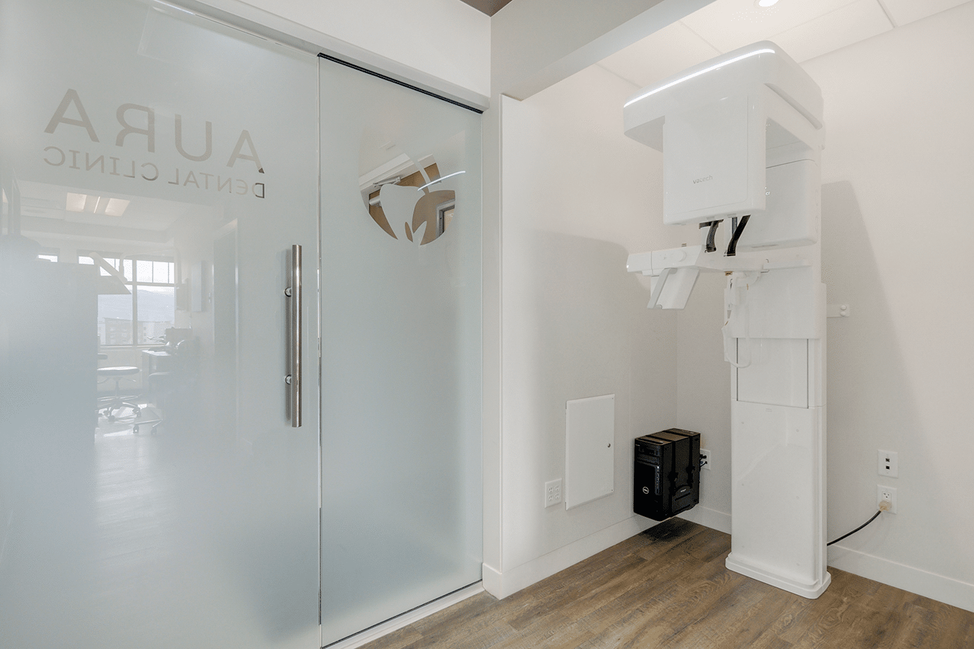 A modern dental clinic entrance with a frosted glass door labeled "AURA Dental Clinic" and a dental X-ray machine nearby.