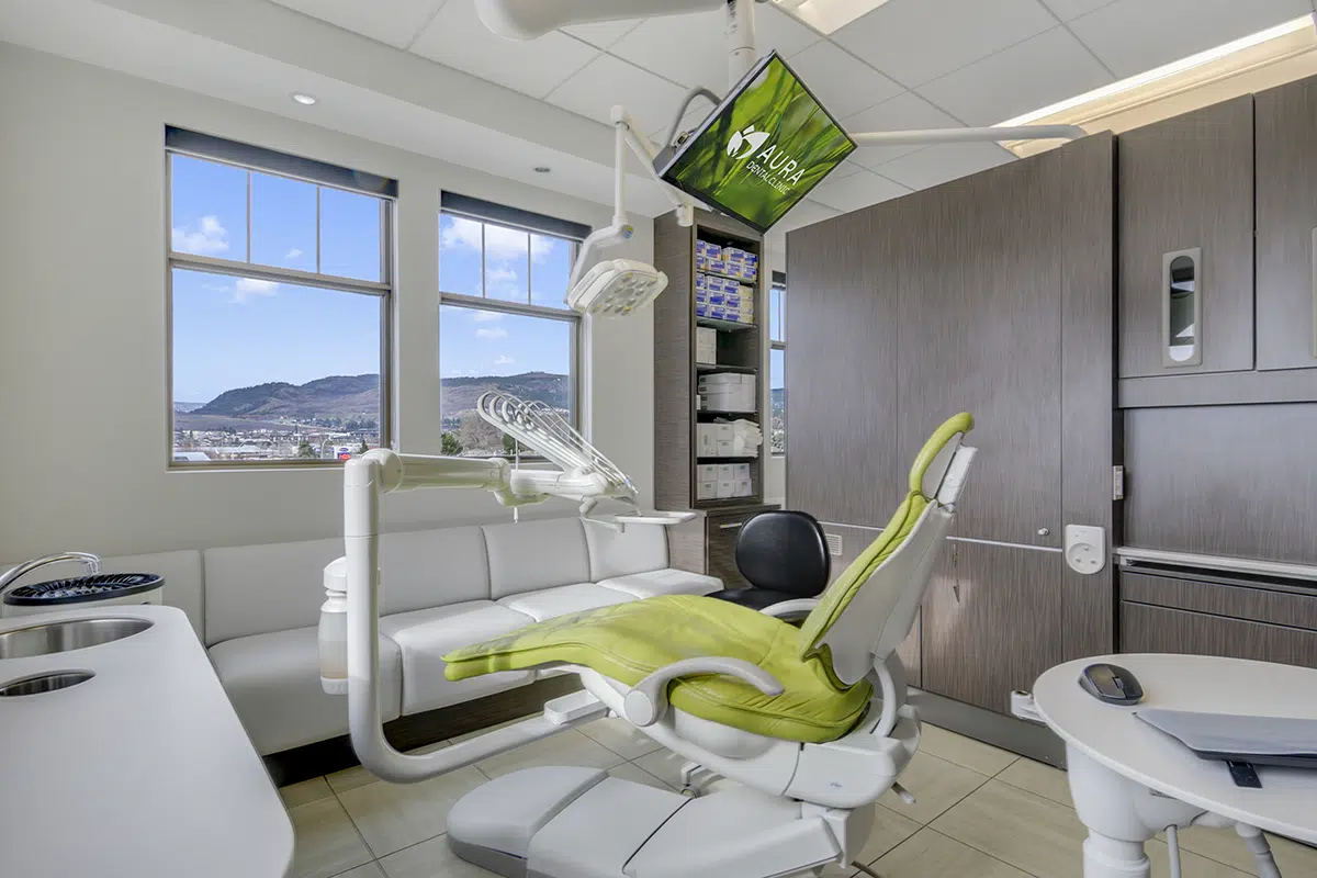 A modern dental office featuring a green dental chair, a monitor displaying "Aura Dental Clinic," large windows with a mountain view, a waiting area with a white sofa, and storage cabinets.