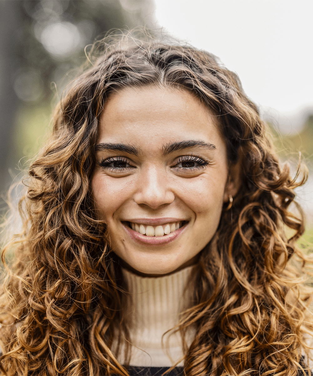 A smiling person with curly hair, wearing a light sweater, outdoors.
