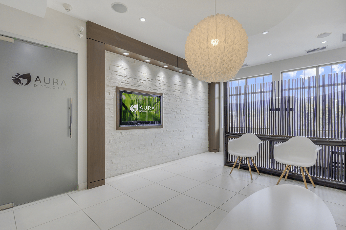 A modern dental clinic reception area featuring a stylish interior design, a logo on the glass door, a wall-mounted TV displaying the clinic's name, and two white chairs.