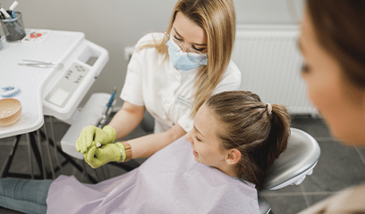 A young girl is sitting in a dental chair, smiling, while a dentist in a mask and gloves is working on her teeth.