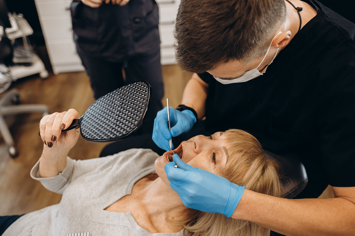 A dentist is examining a patient's mouth while the patient reclines in a dental chair. The dentist is wearing blue gloves and using dental tools, while the patient holds a dental mirror.