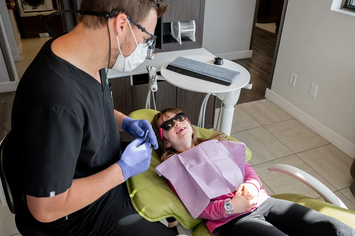 A dentist in scrubs is working on a young girl who is seated in a dental chair, wearing sunglasses and a bib. The dental office has a modern design.