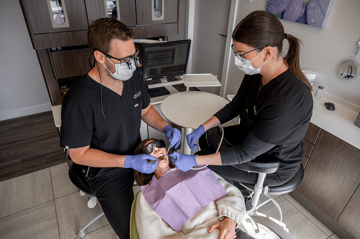 A dental procedure is taking place, with a patient seated and two dental professionals wearing masks and gloves working on the patient's mouth.