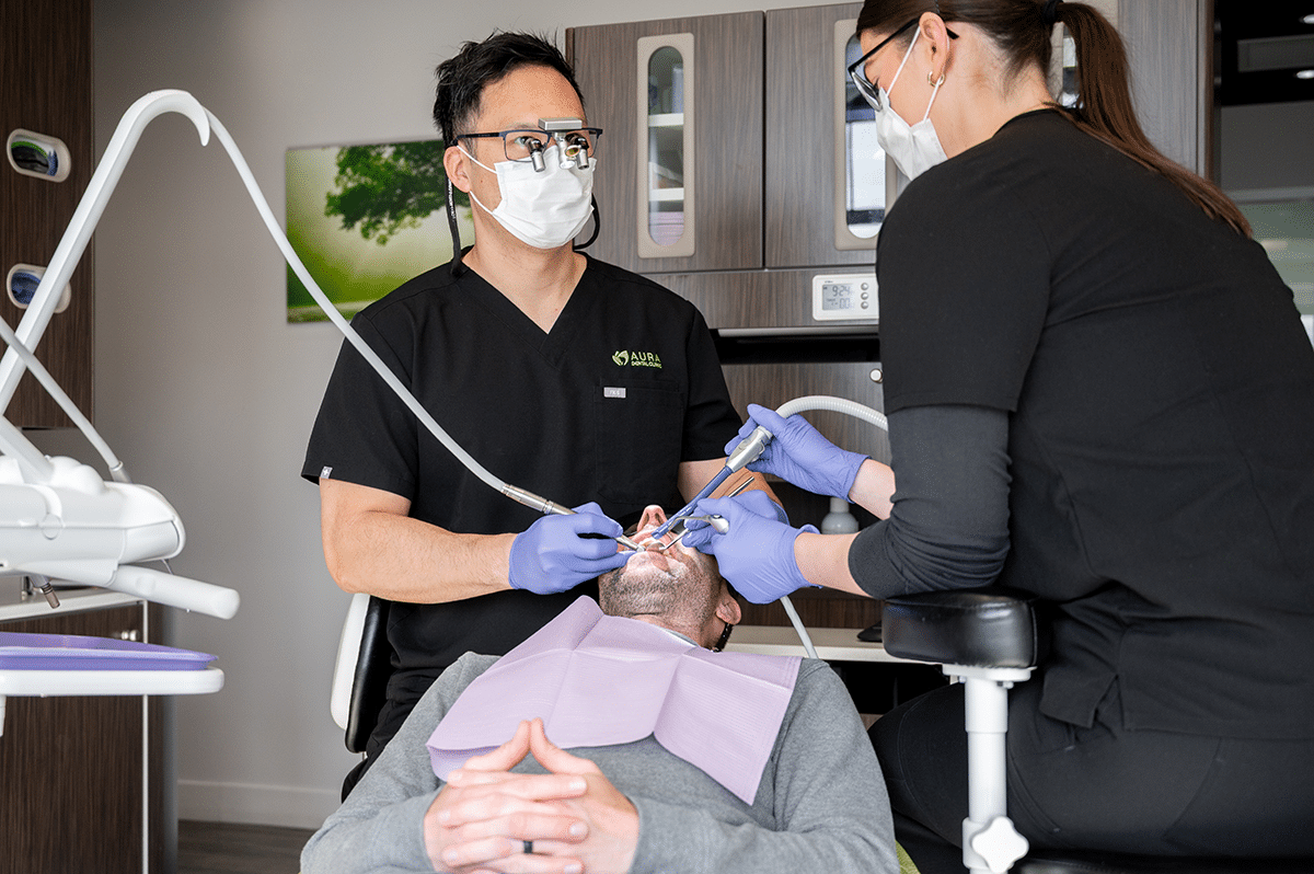 A dentist and an assistant are performing dental treatment on a patient who is reclining in a chair, wearing a bib.