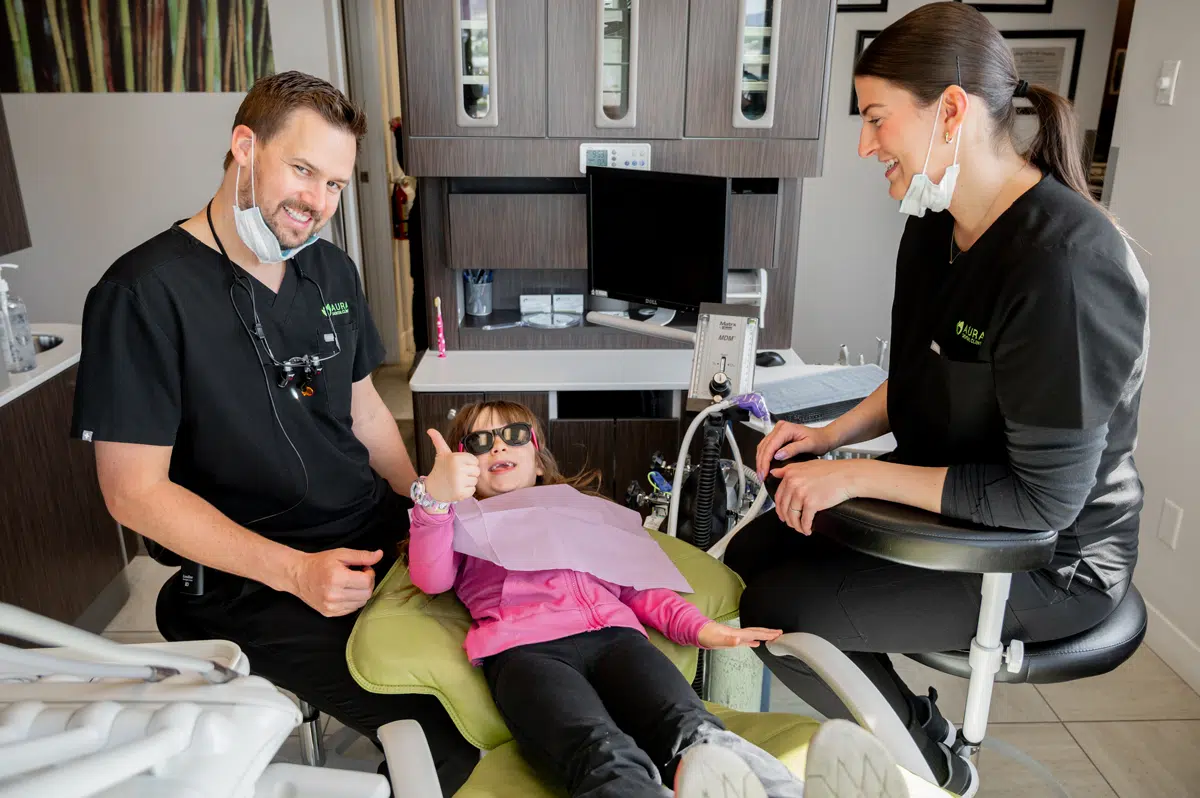A young girl in sunglasses is sitting in a dental chair, smiling and giving a thumbs-up.