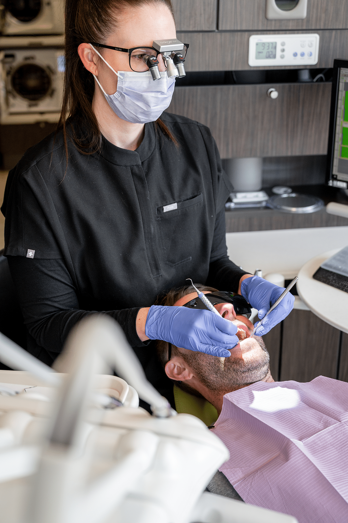 A dental professional in gloves and a mask is using a dental instrument on a patient seated in a dental chair, who is wearing protective glasses.