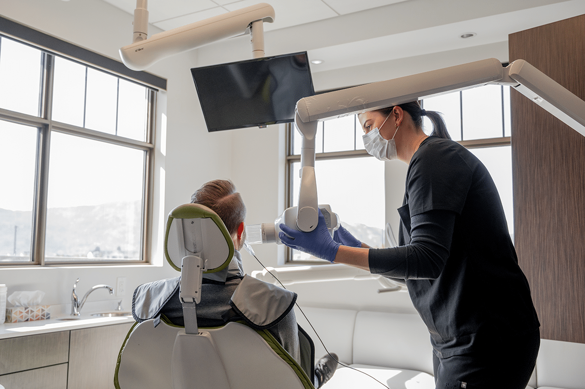 A dental professional wearing gloves and a mask is preparing to use a dental instrument on a patient seated in a dental chair.