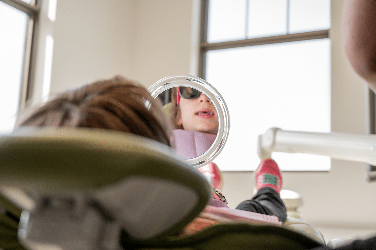 A child wearing sunglasses is reclining in a dental chair, looking into a mirror.
