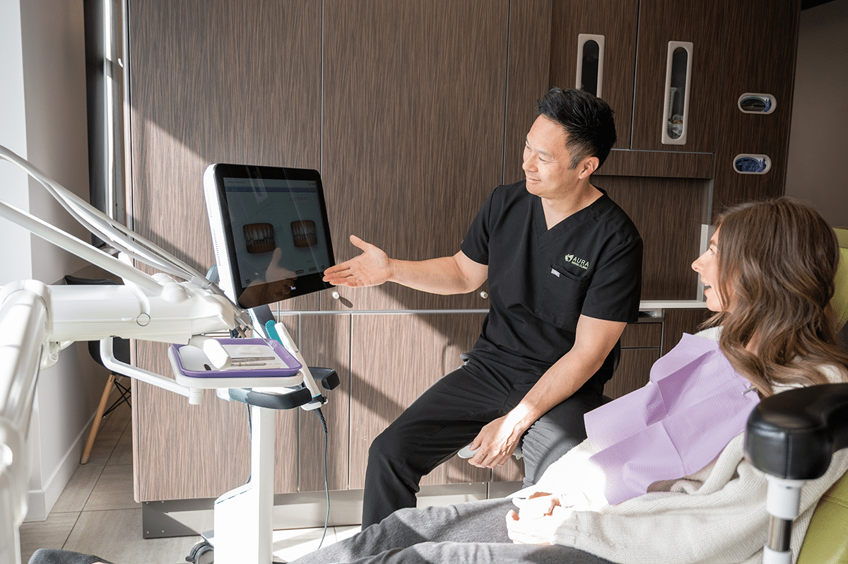 A dentist is showing a patient images on a computer screen in a dental office. The patient is wearing a dental bib and appears engaged in the discussion.