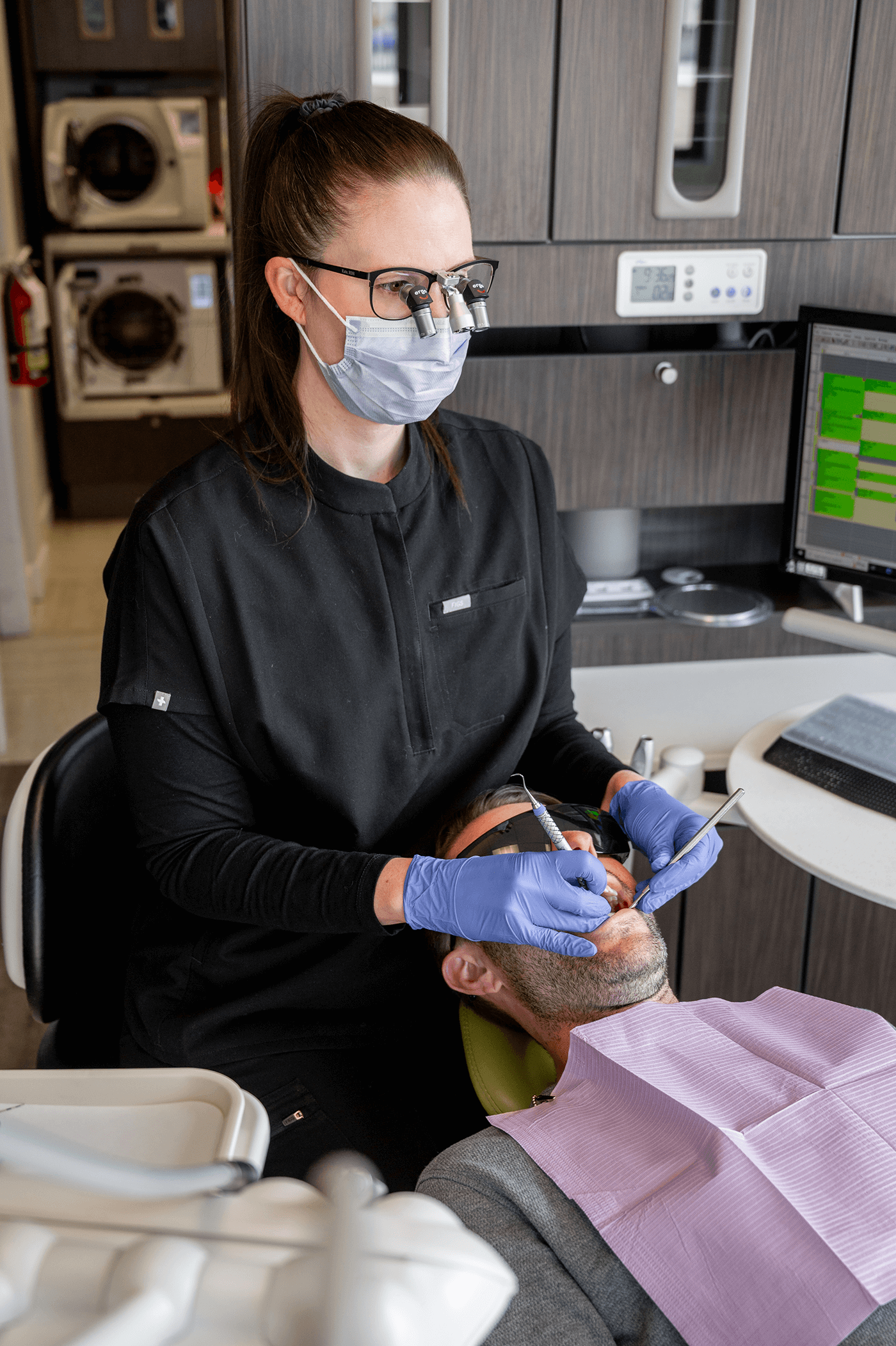 A dental professional wearing a mask and glasses is performing dental work on a patient who is lying in a dental chair with a purple bib.